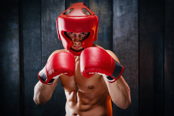 Portrait of sportive muscular athletic man in boxer gloves Dynamic motion portrait of sportive muscular athletic man in boxer gloves, helmet and shorts practicing attack. Fit strength male fighter in movement engaged in training gym, in a fight without rules. headwear stock pictures, royalty-free photos & images