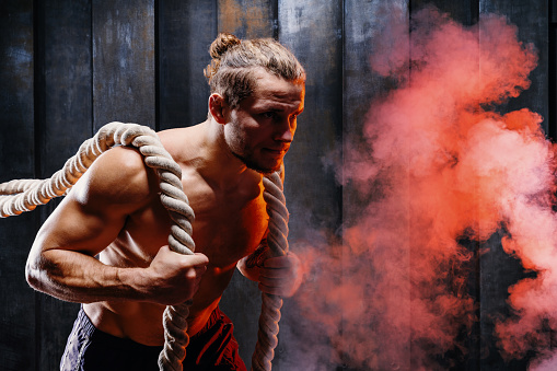 Dynamic motion of muscular man, athlete do exercise with battle ropes. Male training upper body work out at hard gym training. Strong bodybuilder pumping up muscles with smoke on background
