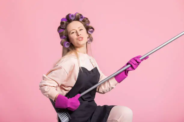 A contented woman happily plays silly while cleaning plays the floor sweeping broom like a guitar. Housewife musically gifted plays house concert on pink background.