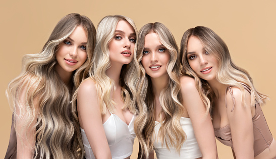 Group portrait of four young  attractive women with exquisite makeup.Elegant long hairstyles dyed in the shades of blonde on them heads. Happy smiles and good mood on the model faces. Hairstyling, dye of hair and makeup.