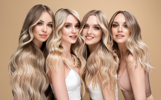 Group portrait of four young  attractive women with exquisite makeup.Elegant long hairstyles dyed in the shades of blonde on them heads. Happy smiles and good mood on the model faces. Hairstyling, dye of hair and makeup.