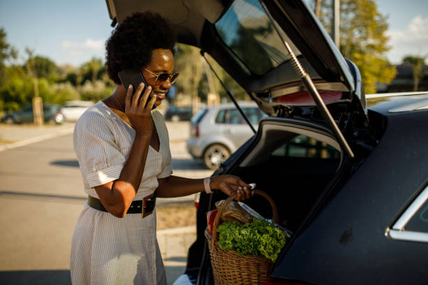 Loading groceries in a car trunk and talking on the phone with a friend Candid shot of smiling mid adult woman returning from groceries shopping, loading a basket in her car trunk and talking on the phone with a friend. curbsidepickup stock pictures, royalty-free photos & images