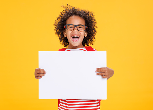 Delighted African American boy in glasses laughing for camera and showing empty poster   while having fun during school  against yellow background