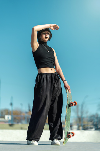 Pretty cool skater girl with eye glasses posing with her skate board towards the sunlight. Vertical photography