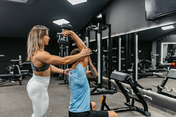 istock Young Female Trainer Helping Young Man With Lifting Weights In The Gym 1423614955