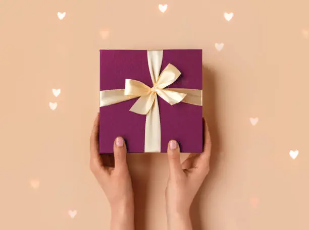 Female hand with natural manicure holding violet gift box with light golden ribbon on trendy beige background. Xmas and New Year postcard design. Black Friday sales, Birthday celebration party concept