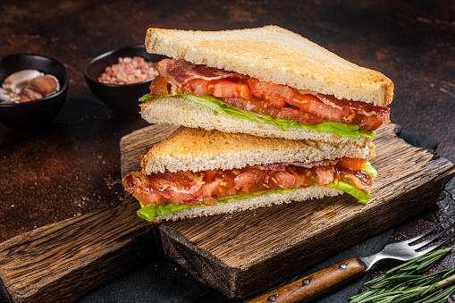 Classic BLT sandwich with bacon, tomato and lettuce on wooden board with herbs. Dark background. Top view.