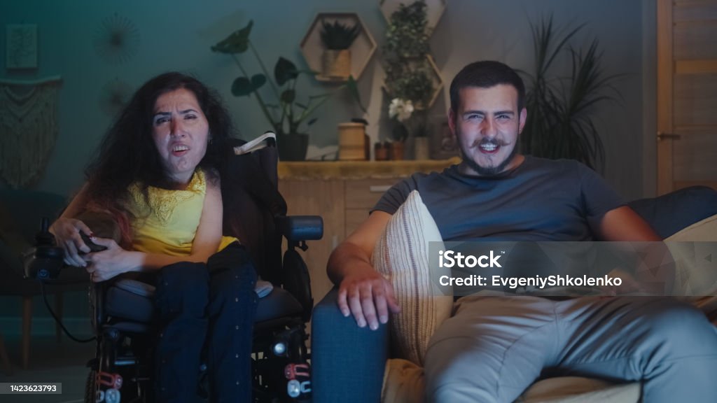 Woman with disability and man watching TV Shocked woman with physical disability in a wheelchair and man wondering and looking at each other, while watching TV show together at home in the evening 20-24 Years Stock Photo