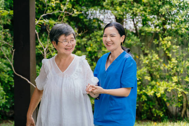 Happy female caregiver in blue scrubs holding hand of senior woman and looking away with smile while walking near lush trees in garden together Happy female caregiver in blue scrubs holding hand of senior woman and looking away with smile while walking near lush trees in garden together walking aide stock pictures, royalty-free photos & images