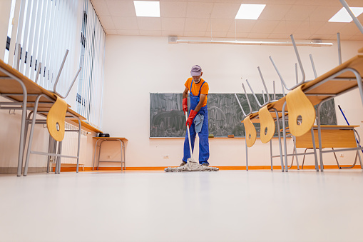 A front view of an unrecognizable mature janitor mopping the floor of the empty school classroom, and disinfecting during Covid-19.