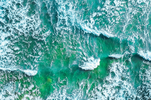 Dramatic seascape seen from above, while breaking waves splash with foam. Blue and jade hues. Aerial view.