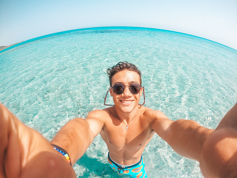 Portrait of happy young man taking a selfie of him in the beach in a blue turquoise water having fun and enjoying alone vacations outdoors.