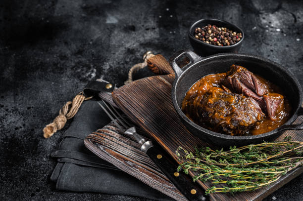istock Stewed beef cheeks with brown red wine sauce in skillet. Black background. Top view. Copy space 1423632440