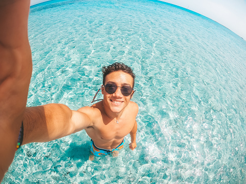 Portrait of happy young man taking a selfie of him in the beach in a blue turquoise water having fun and enjoying alone vacations outdoors.