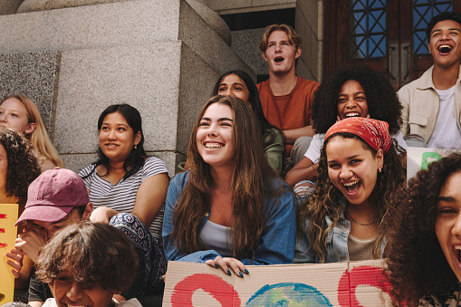 Group of youth activists smiling happily while sitting outside a building with posters and banners. Multicultural young people protesting against global warming and climate change.