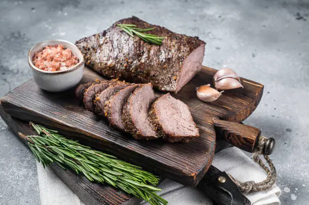Roast and sliced tri tip beef steak on a wooden board with herbs. Gray background. Top view.