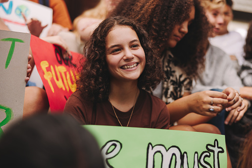 Happy teenage girl smiling cheerfully while sitting with a group of climate activists. Multicultural protestors striking for climate justice and environmental sustainability.