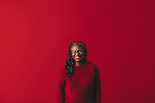 Black woman with dreadlocks laughing happily while standing against a red background. Cheerful mature woman embracing her natural hair with pride.