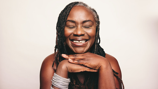 Woman with dreadlocks smiling with her eyes closed against a studio background. Happy mature woman wearing light make-up and jewellery. Cheerful middle-aged woman embracing her ageing body.