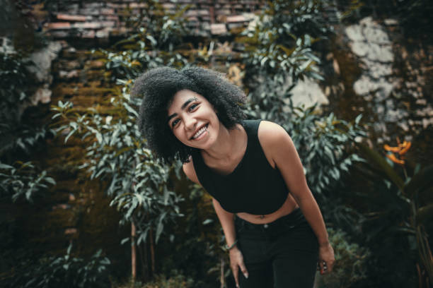 Beautiful young mixed-race woman, wearing black crop top, with afro hair and piercings, surrounded by greenery, posing next to rustic wall at Tay Ho, Hanoi, Vietnam
