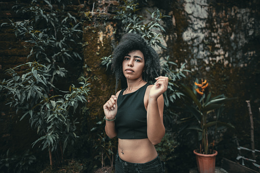 Beautiful young mixed-race woman, wearing black crop top, with afro hair and piercings, pooling hair, surrounded by greenery, posing next to rustic wall at Tay Ho, Hanoi, Vietnam