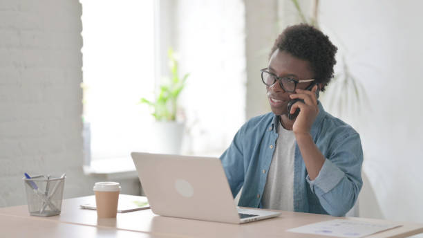 istock Young African Man Talking on Phone while using Laptop in Office 1423646162