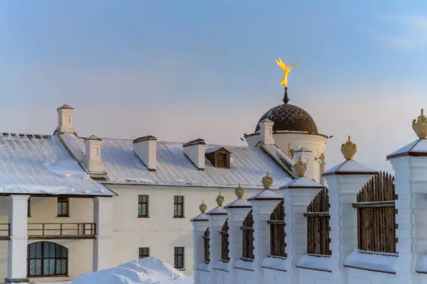 A golden angel plays the trumpet on one of the towers of the Tobolsk Kremlin (Russia) against the blue morning sky. Winter with snowdrifts. Ancient buildings and a fence with white embossed pillars