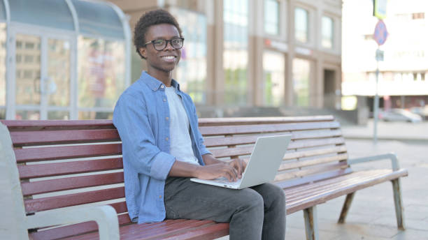 istock Young African Man with Laptop Smiling at Camera while Sitting Outdoor on Bench 1423648769