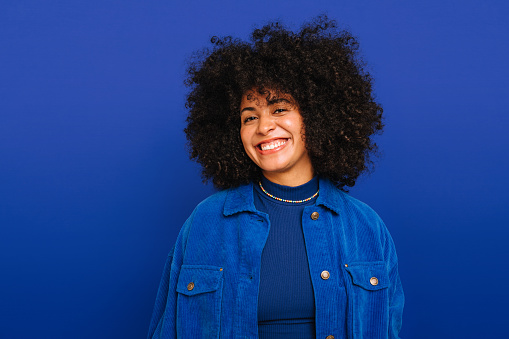 Feeling good in blue. Happy woman with curly hair smiling at the camera while standing against a blue background. Beautiful woman with curly hair wearing her natural air with pride.