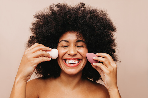 Happy young woman smiling cheerfully while holding two make up sponges on her face. Gorgeous woman with Afro hair using beauty blenders to apply cosmetic foundation on her face.