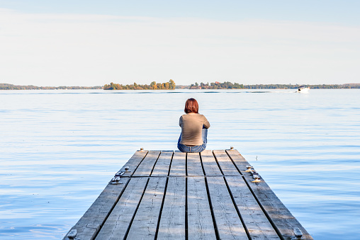 Woman sitting alone at the end of a wooden jetty on the bank of a large river on a sunny autumn day. River St. Lawrence, Thousand Islands National Park, ON, Canada.
