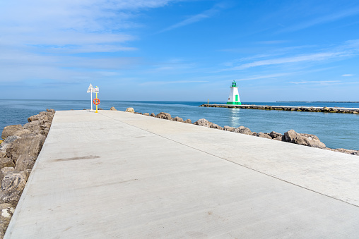 Concrete footpath on the breakwater at the entrance of a harbour on a lake. A lighthouse is on the breakwater at the other side of the canal. Lake Ontario, ON, Canada.