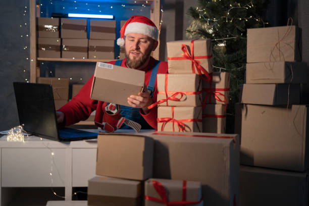 święty mikołaj w kapeluszu trzymający pudełko z prezentami za pomocą laptopa siedzącego przy stole w domowym biurze późno w wigilię bożego narodzenia. witryna e-commerce święta święta zakupy online sprzedaż e-commerce - santa claus christmas watch workshop zdjęcia i obrazy z banku zdjęć