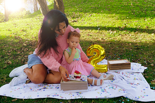interracial family of Venezuelan mother celebrating daughter's second birthday in the park during a picnic with balloons and strawberry cake