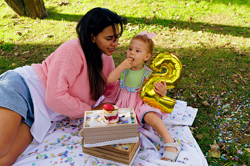 little girl celebrating her second birthday holding a balloon next to her Latina mother in a park under a tree during a picnic.