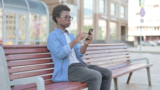 istock Young African Man Browsing Internet on Smartphone while Sitting Outdoor on Bench 1423677974