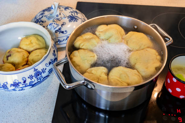 Sweet fruit dumplings floating in boiling water in pot on hot ceramic hob, boiled dumpling in decorative bowl, lid, melted butter in small red dotted pot. Boiling dumpling on hob.