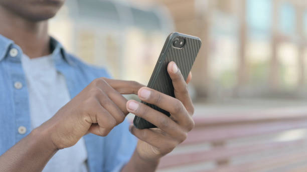 istock Close Up of Young African Man Using Smartphone while Sitting Outdoor on Bench 1423680437