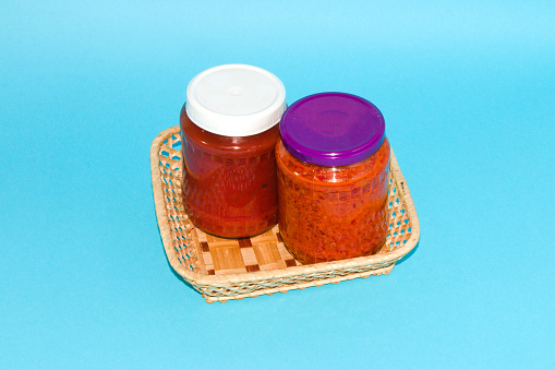 two jars of ajvar in a wooden basket on a blue background, ajvar traditional balkan paprika dish