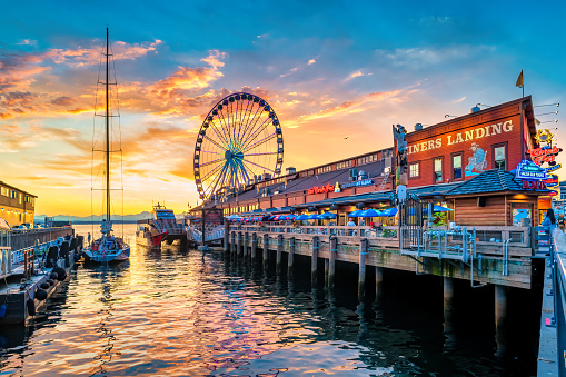 People walk and dine at the downtown Seattle waterfront with the landmark Great Wheel in the background, at sunset time.