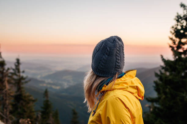 Woman with knit hat and yellow jacket looking at mountain range during sunset stock photo
