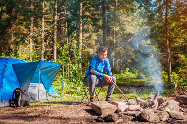 istock Man traveler relaxing by campfire in forest sitting next to tent checking fire. Summer camping. Traveling alone 1423703484