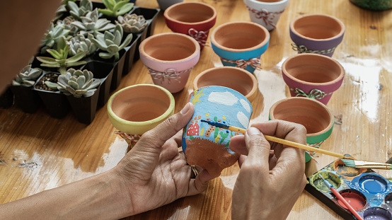 Hands of a Latin mulatto woman, painting clay pots to plant succulent plants on a wooden table with natural lighting and acrylic paints