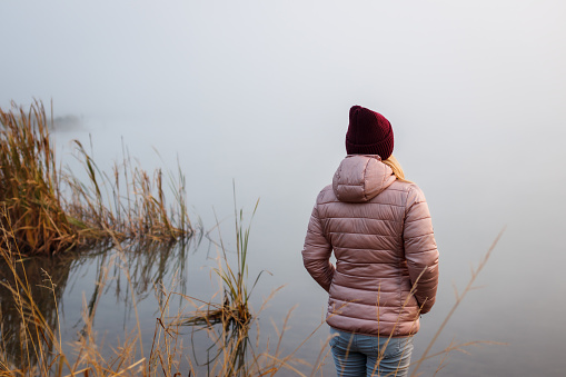 Woman standing by lake. Enjoyment and feeling harmony with a nature. Misty morning at cold weather