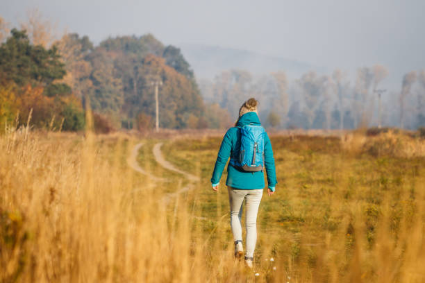 Woman with backpack hiking on footpath at cold morning. Fall exploration outdoors in rural scenery