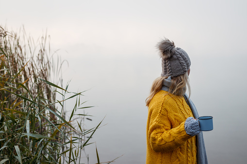 Woman relaxing with hot drink next to lake at autumn cold morning. Camping outdoors at fall season. Blond hair woman wearing knit hat and yellow sweater