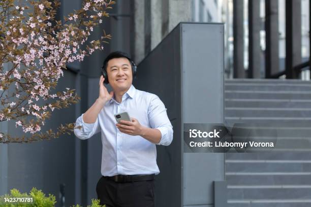 Satisfied Asian Man In White Shirt Holds Phone And Listens To Audiobook Podcast In Black Headphones Stock Photo - Download Image Now