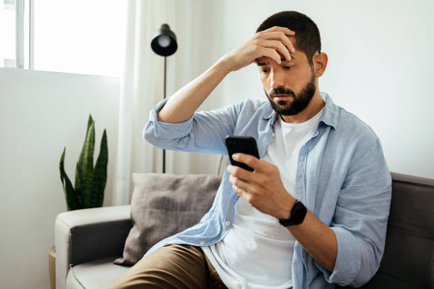 hombre triste revisando el teléfono inteligente sentado en un sofá en casa - worried fotografías e imágenes de stock