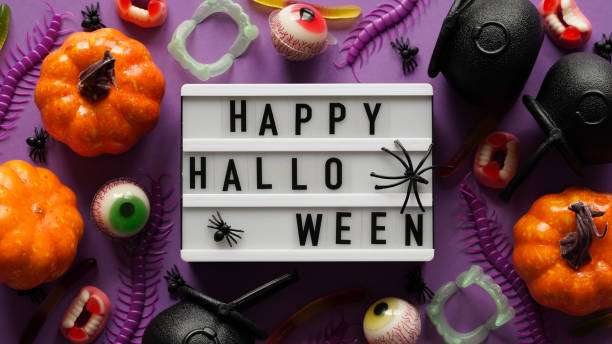istock Pumpkins, cauldrons, pots, worms, jaws, spiders, eye-shaped candies and text board with the inscription Happy Halloween on a violet background, top view. A collection of items for a Halloween party. 1423720006