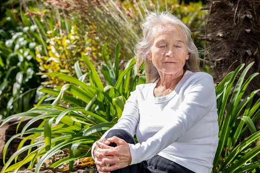 Portrait of modern senior woman sitting in the garden full of sunshine with her eyes closed, background with copy space, full frame horizontal composition